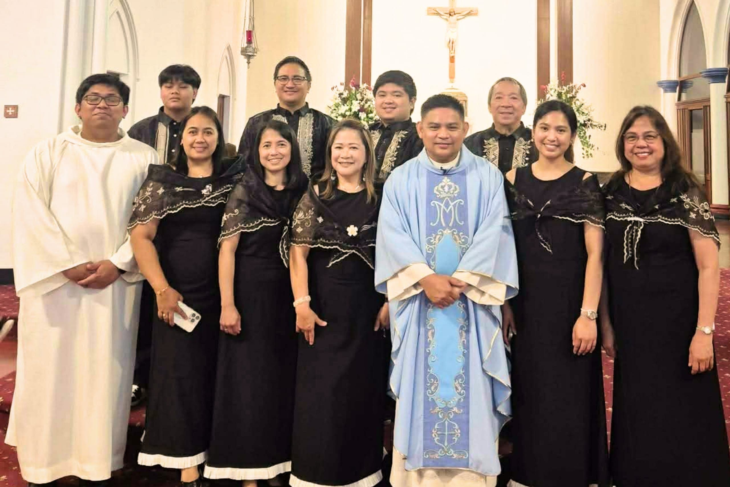 Choir members in black Filipiniana dresses standing with a priest and altar server in the sanctuary